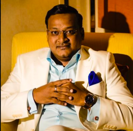 Man wearing glasses, a light blue shirt, and a white blazer with a blue pocket square, sitting with hands clasped.